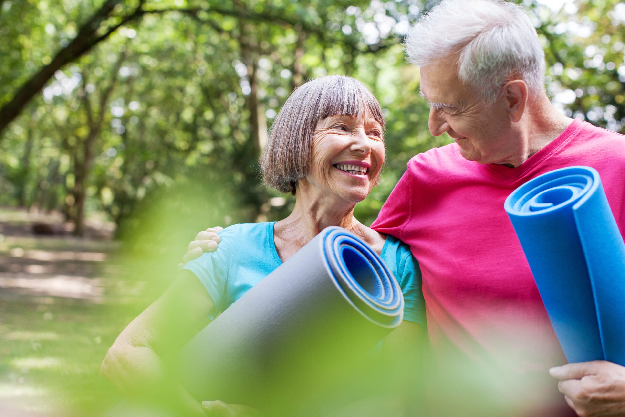 A smiling couple stands together in the park with yoga mats